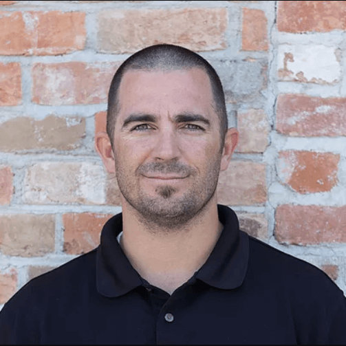 A physical therapist named Trent Henderson, wearing a black shirt, stands in front of a brick wall.